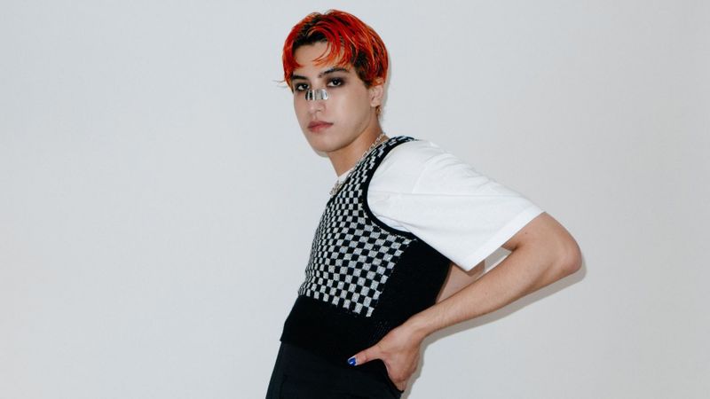 White/gray background. Man with wet red hair wearing heavy black eyeliner and nose bandaid. 