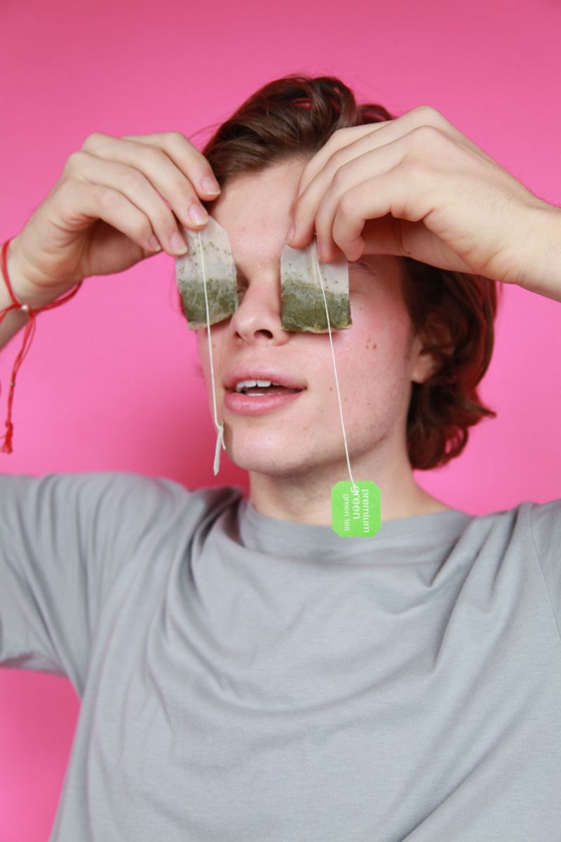 Pink background. Guy holding teabags on top of this eyes. 